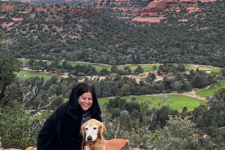 Woman with a dog on a rocky overlook, red cliffs and greenery in the background.