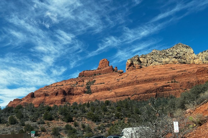 Red rock formations under blue sky with wispy clouds, vehicles on road at base.