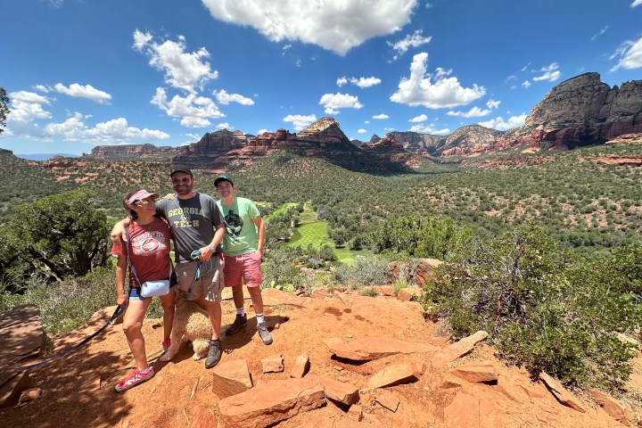 Three people smiling on a rocky hilltop with a scenic desert landscape in the background.