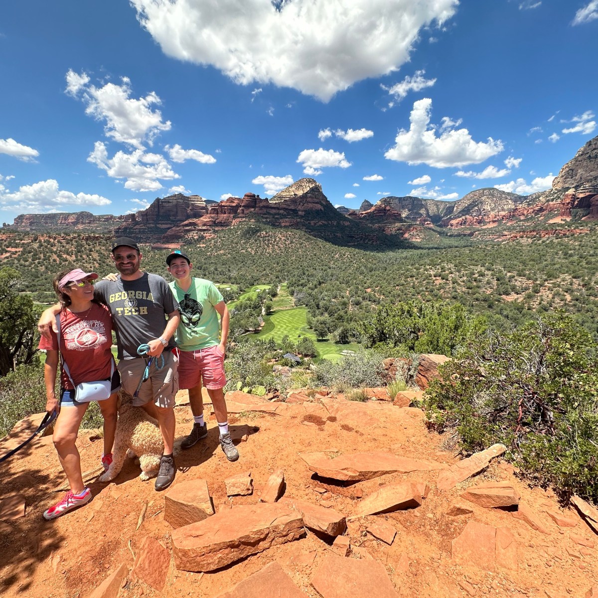 Three people smiling on a rocky hilltop with a scenic desert landscape in the background.