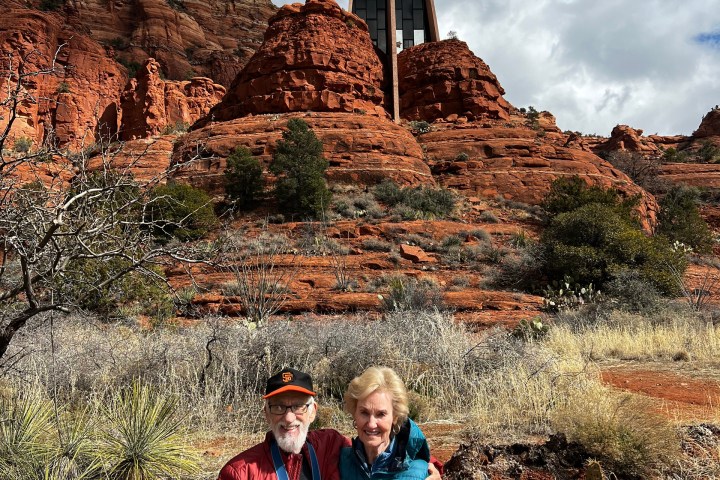 Two people pose in front of rock formations and a modern building under a cloudy sky.