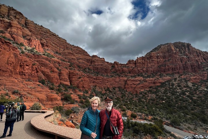 A couple stands on a walkway with a backdrop of red rock cliffs and cloudy sky.