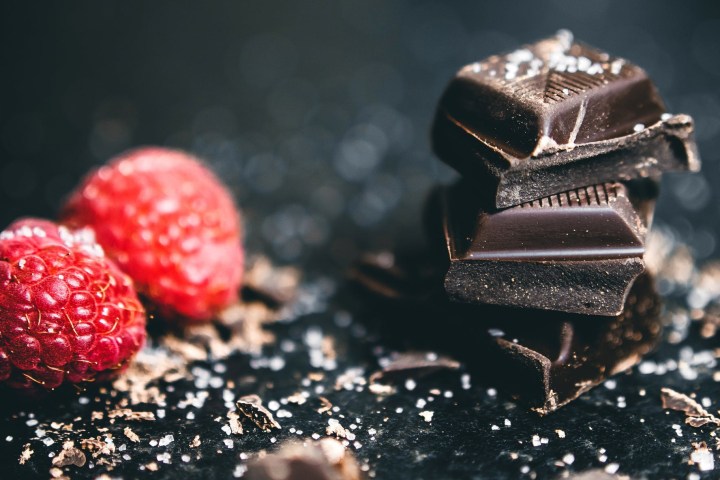 Two raspberries next to stacked chocolate pieces on dark surface.