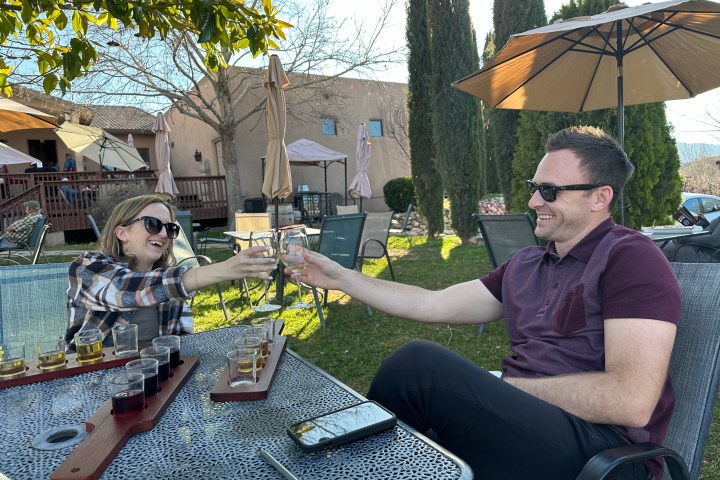 Two people clinking glasses outdoors, seated at a table with flights of drinks.
