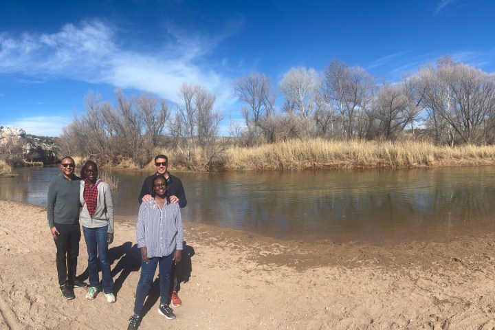 Four people standing by a riverbank under a clear blue sky with trees in the background.