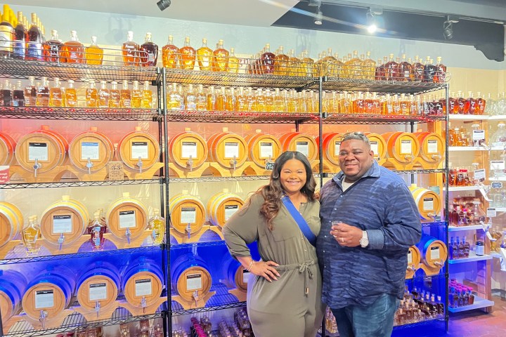 Two smiling people stand in front of shelves with bottles and barrels.