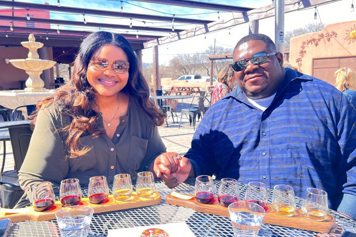 Two people sitting at an outdoor table with sunglasses, holding hands, and enjoying a drink tasting.