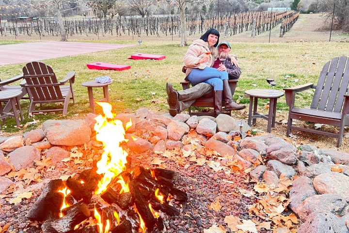 Couple sitting by a fire pit with vineyard in the background and empty chairs nearby.