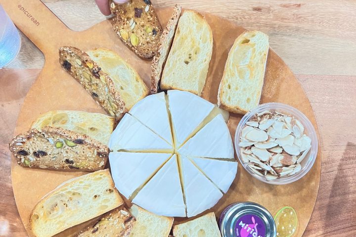 Cheese board with sliced cheese, bread, nuts, jam, and a hand reaching for a piece, on a wooden table.