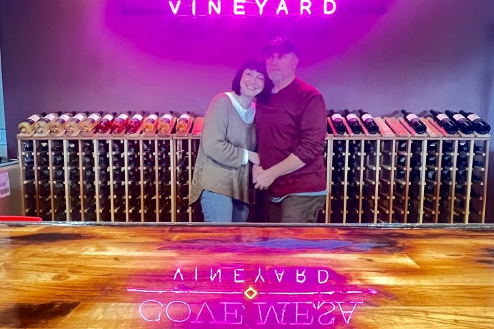 Couple standing in front of neon sign 'Cove Mesa Vineyard' with wine bottles on racks behind them.