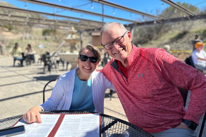 Two smiling people sitting outdoors at a table with menus, under a pergola, with a sunny background.
