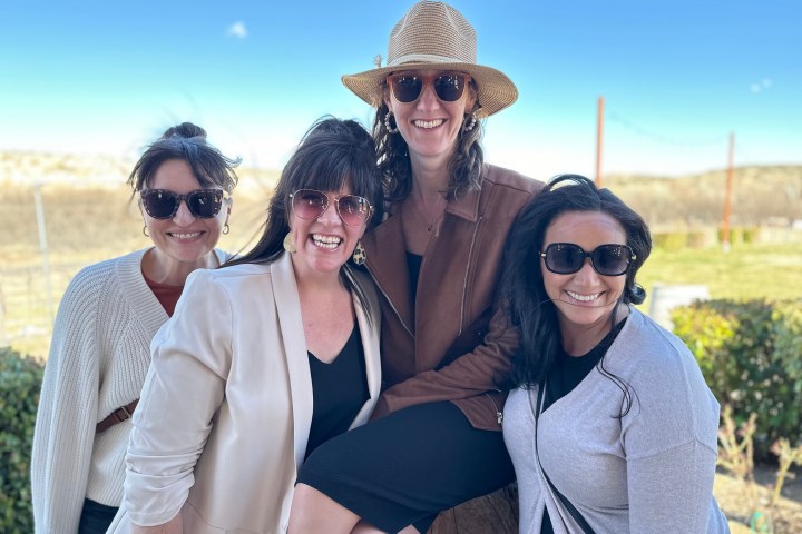 Four women wearing sunglasses smiling outdoors on a sunny day.