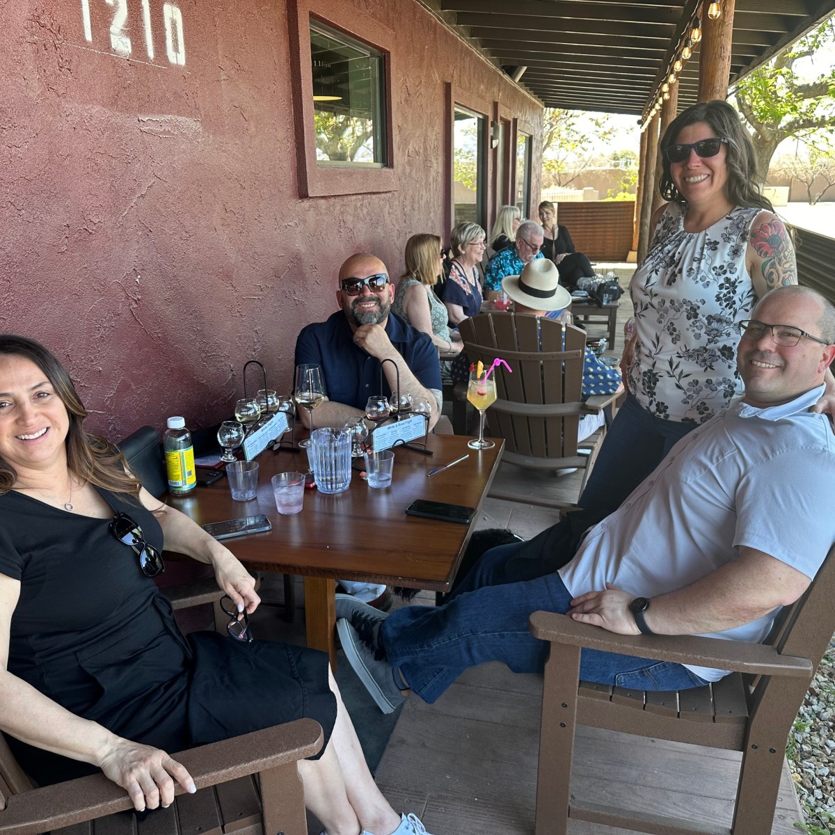 Group of people sitting and smiling on a patio with drinks on a wooden table.
