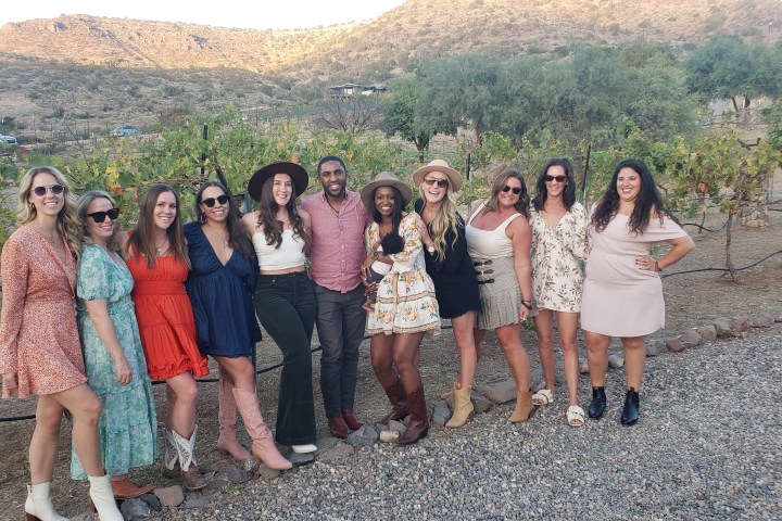 Group of people posing outdoors in casual attire with hills and vineyard backdrop.