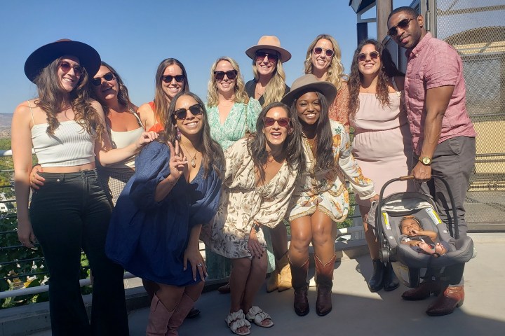 Group of people smiling together outdoors, some wearing hats and sunglasses, with a baby in a carrier on the right.