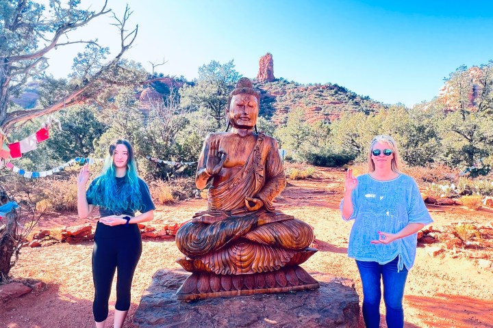 Two people standing beside a wooden Buddha statue in a desert landscape with prayer flags.