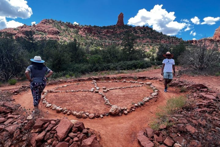 Two people standing near stone circle on red desert path, with rocky hills and blue sky in the background.