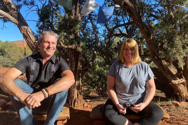 Two people sitting outdoors on wooden benches under trees with colorful flags, in a sunny setting.