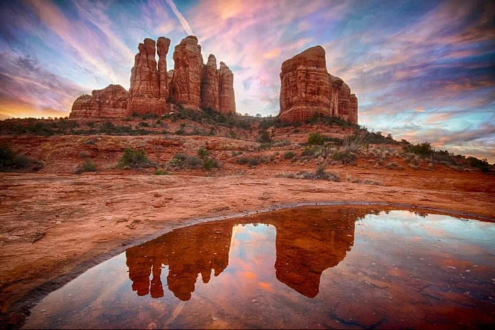 Red rock formations under a colorful sky, reflected in a small pool of water.