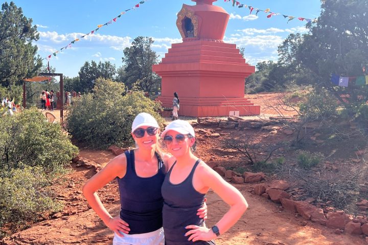 Two people standing in front of a red stupa with prayer flags under a bright blue sky.