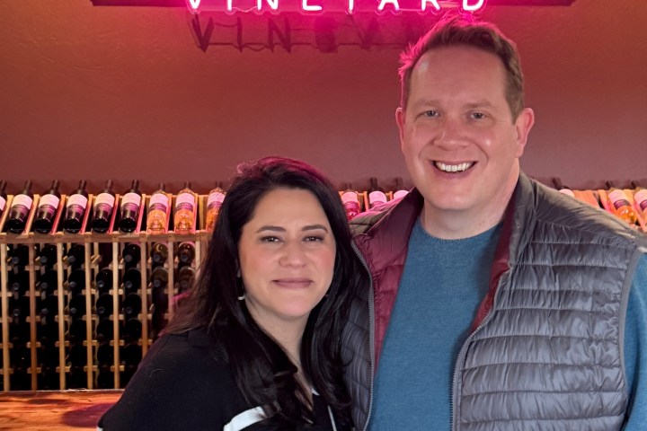 Two people smiling in front of a neon sign reading 'Cove Mesa Vineyard' with wine bottles visible behind them.