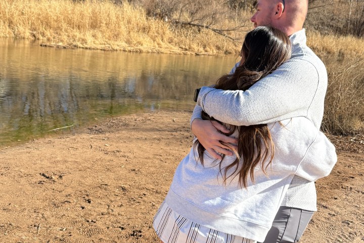A couple embracing by a riverside with bare trees and a clear blue sky.