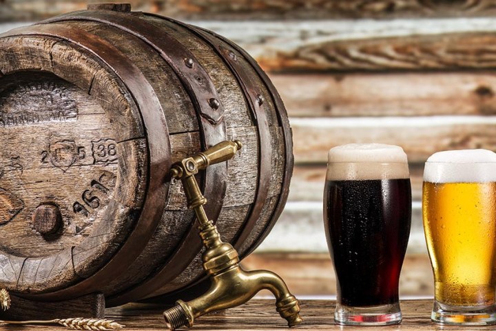 Two beer glasses next to a wooden barrel with a tap, on a rustic wooden background.