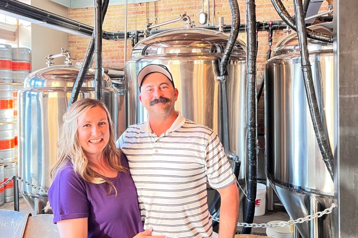 Two people smiling in front of large stainless steel brewing tanks in a brewery.