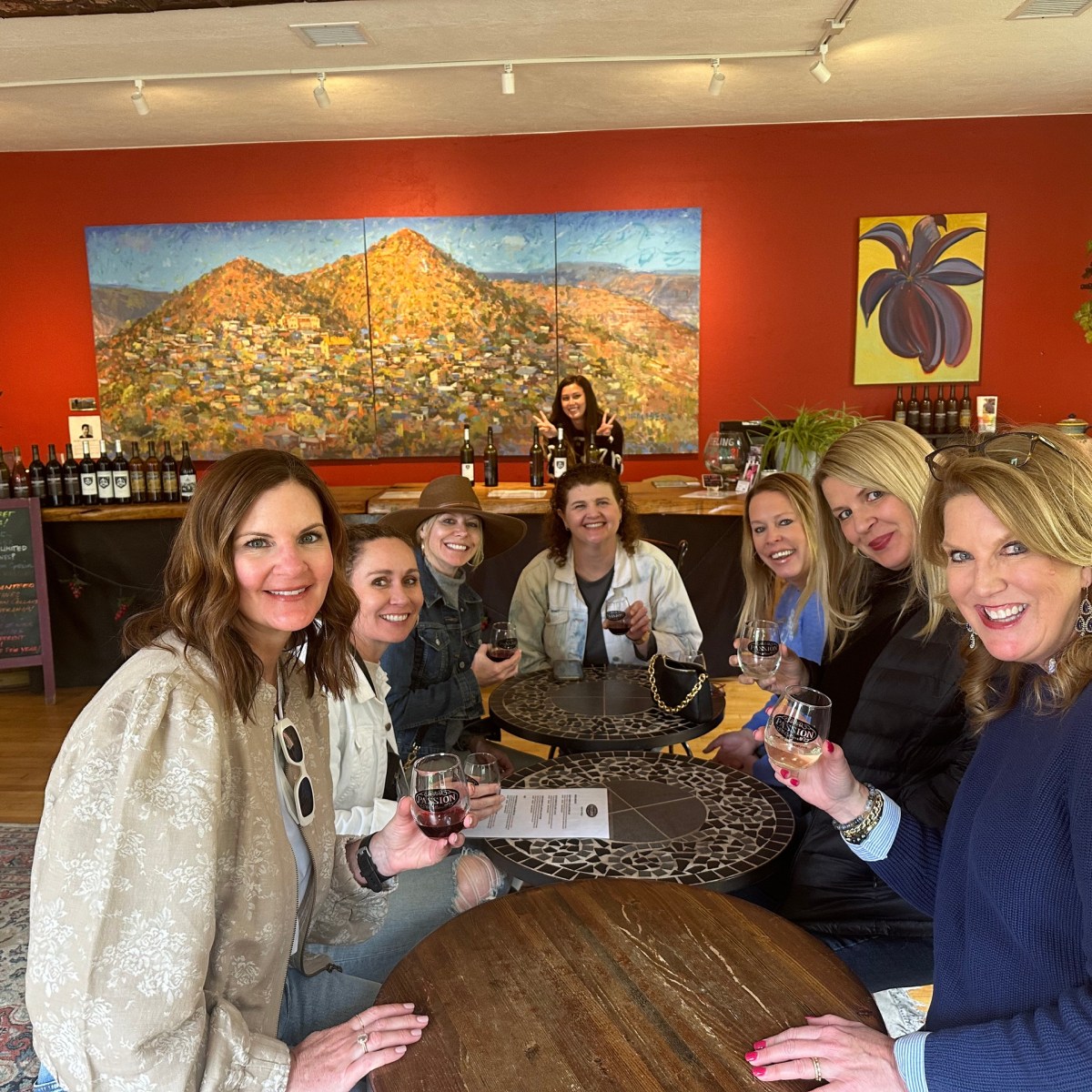 Group of women sitting indoors, holding wine glasses, with colorful wall art in the background.
