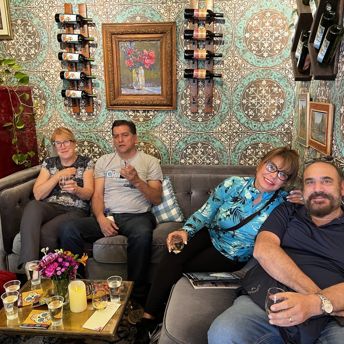 Four people sitting on a couch with drinks, surrounded by art on patterned wallpaper.