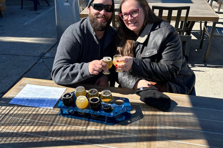Two people sitting outdoors at a table, holding small glasses, with a tray of drinks in front.