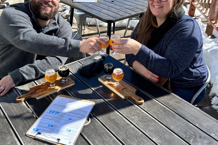 Two people clinking beer glasses at an outdoor table with snow around.