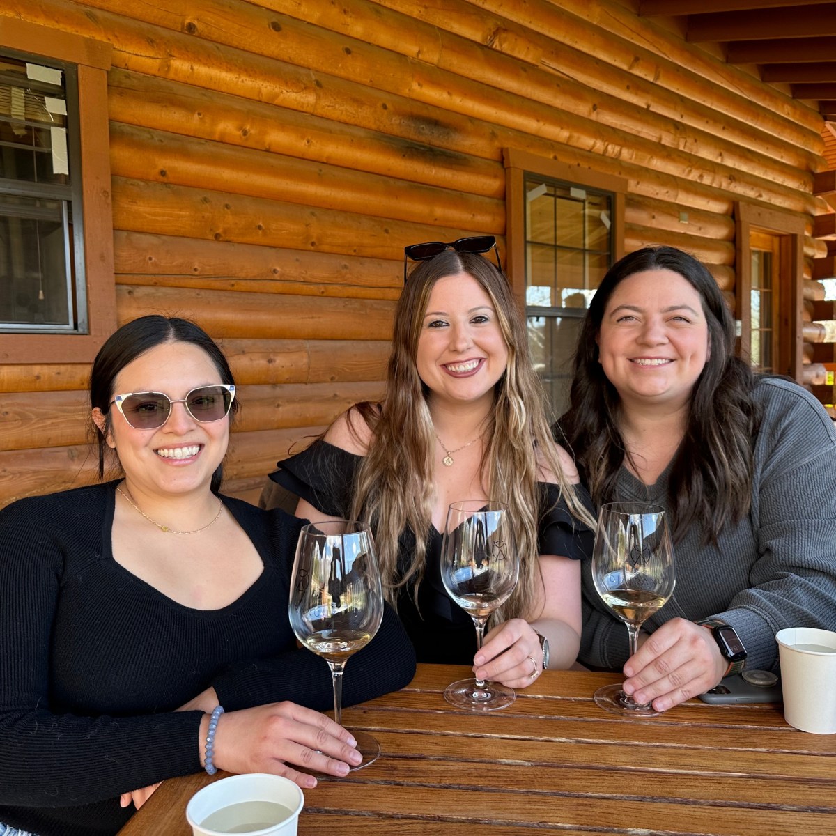 Three women smiling and holding wine glasses at a wooden table on a lodge deck.