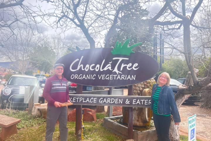 Two people stand beside a sign for ChocolaTree Organic Vegetarian under overcast skies.
