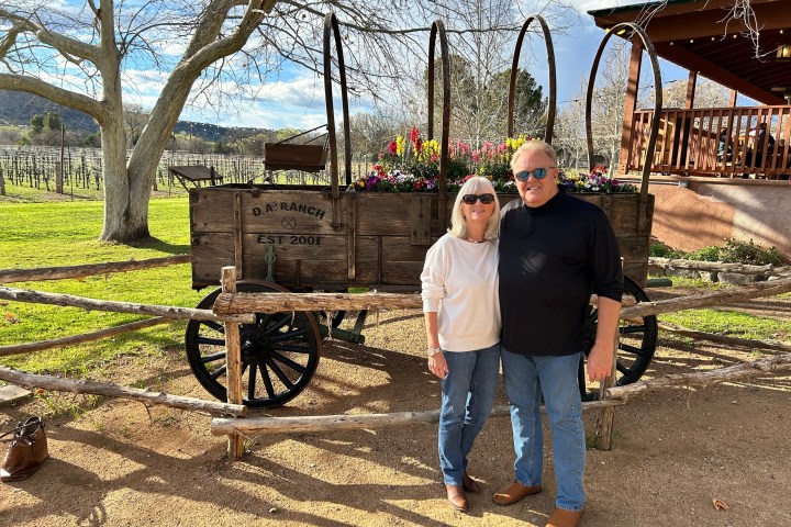Couple standing in front of a vintage wooden wagon with flowers, under a large tree on a sunny day.
