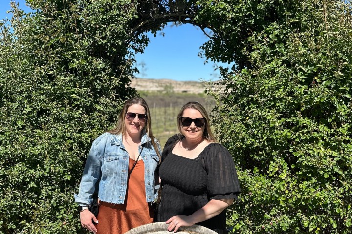 Two women posing by a wine barrel under a leafy archway in a vineyard.