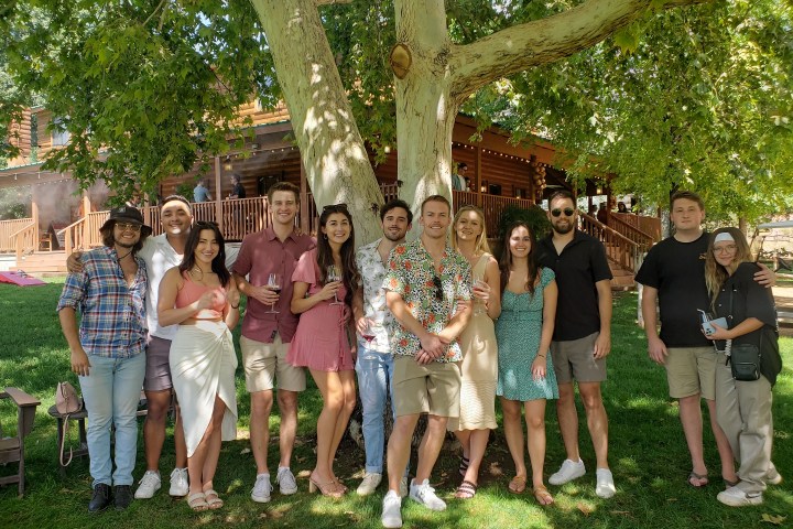 A group of people smiling and posing under a large tree in front of a wooden house.