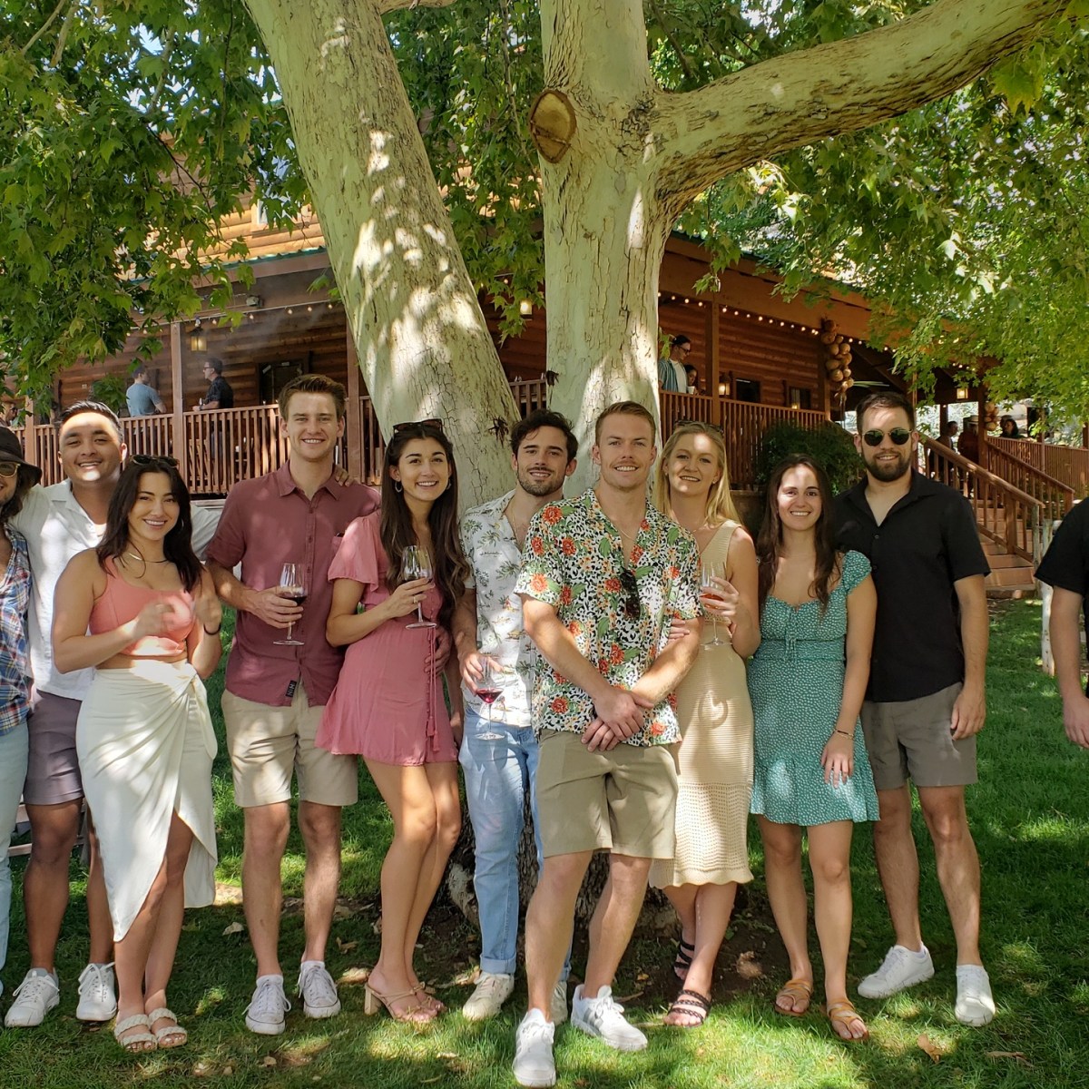 A group of people smiling and posing under a large tree in front of a wooden house.