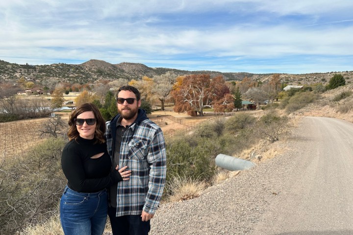 Couple in sunglasses poses on a rural road with hills and trees in the background.
