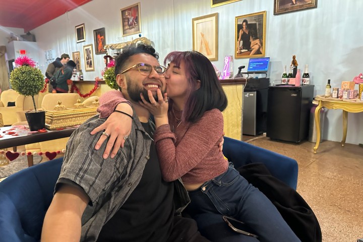 A woman kisses a smiling man on the cheek in a colorful, decorated room with framed photos on the wall.