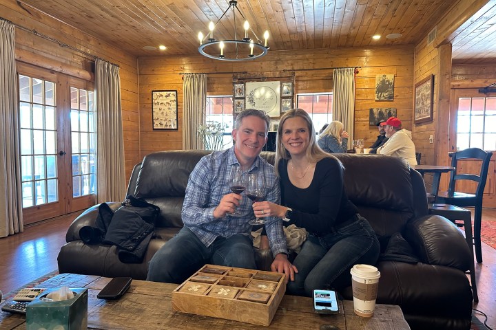 Couple sitting on a leather couch in a rustic room, holding wine glasses, smiling at the camera.