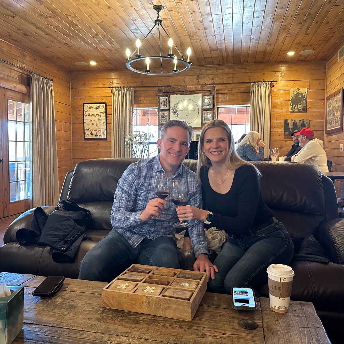 Couple sitting on a leather couch in a rustic room, holding wine glasses, smiling at the camera.