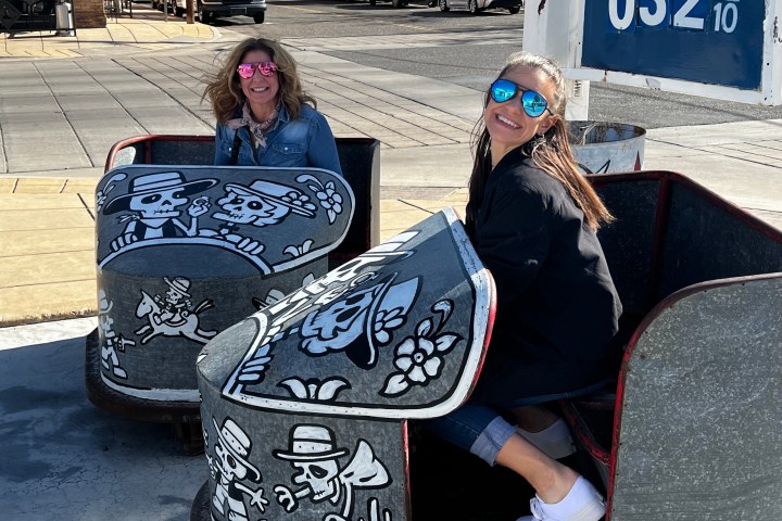 Two women in sunglasses sit in decorated metal carts with skeleton art on a sunny street.