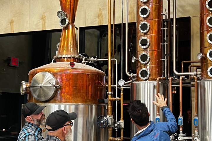 Three men examining a large copper and stainless steel distillation apparatus indoors.