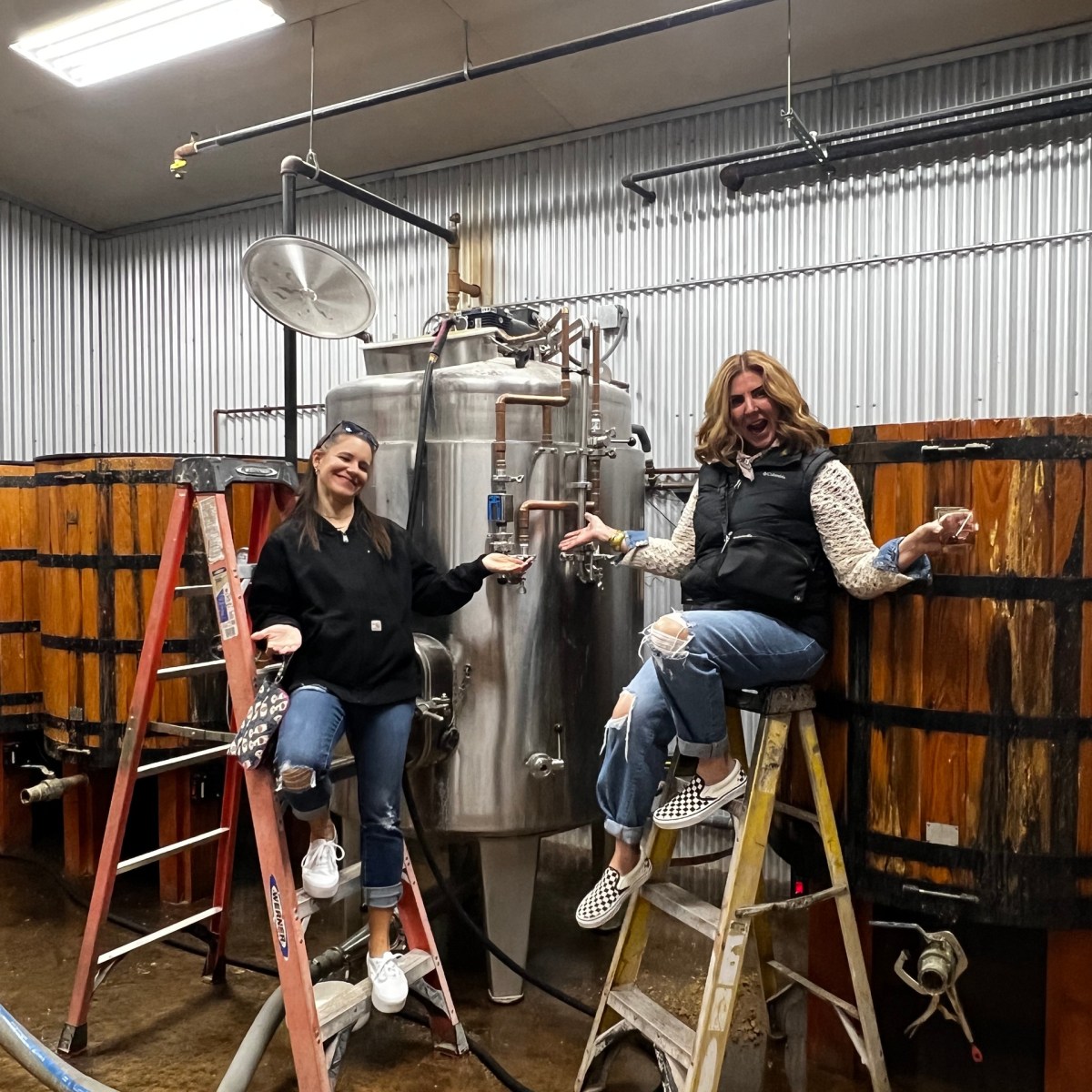 Two people on ladders in a distillery, surrounded by wooden barrels and metal tanks.
