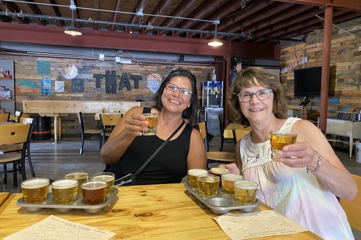 Two women smiling and holding beer flights in a rustic pub setting.