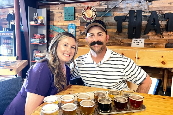 Smiling couple seated at a wooden table with beer flights in a cozy brewpub setting.