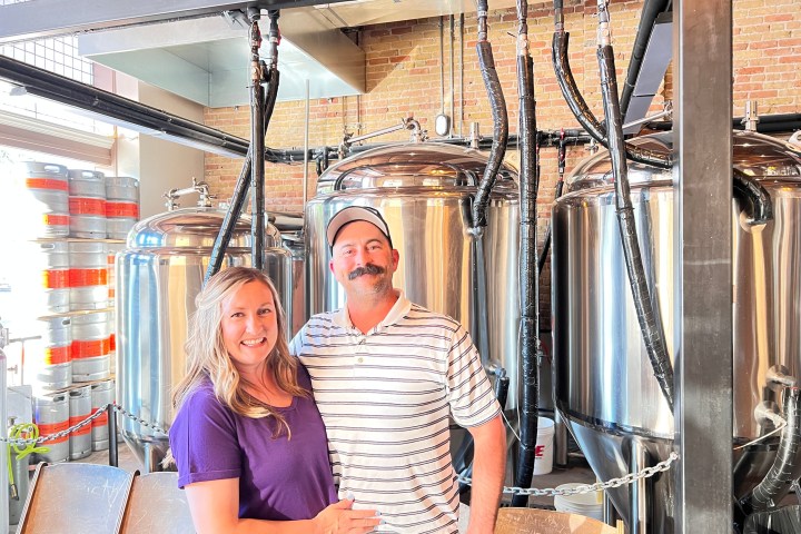 Two people smile in front of large metal brewing tanks inside a brewery.