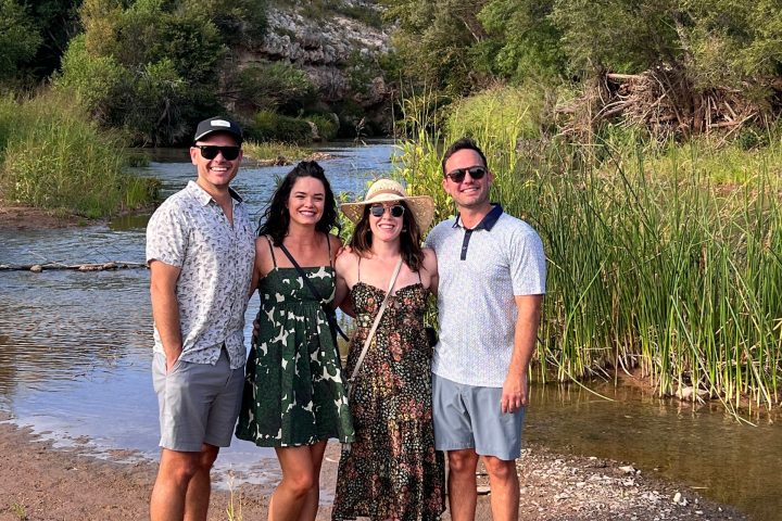 Four people standing by a river with grass and trees in the background.
