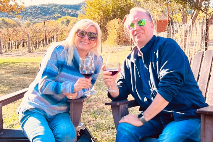 Two people sitting outside with wine glasses, enjoying a sunny day in a vineyard.